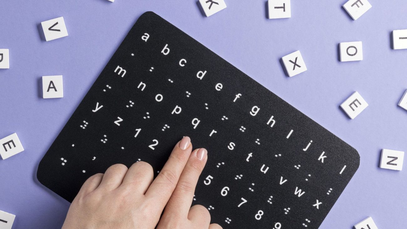 Manos leyendo alfabeto braille en tableta táctil negra con letras y números, rodeada de fichas de Scrabble con letras del alfabeto regular
